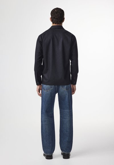 En person står vänd bort, iklädd en svartrandig skjorta och blå vida jeans, framför en enfärgad vit bakgrund.