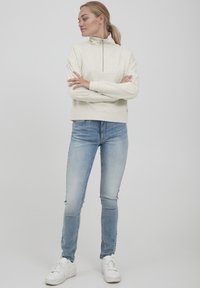 Lys beige fleece-pullover med høj krave og lynlås, parret med lyseblå slim-fit jeans og hvide sneakers. Simpelt design og tekstur.