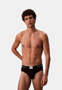 Schwarzer Baumwoll-Slip mit weißem elastischem Bund, der das Calvin Klein-Logo trägt, glatter Textur und körperbetontem Schnitt. Model posiert lässig.