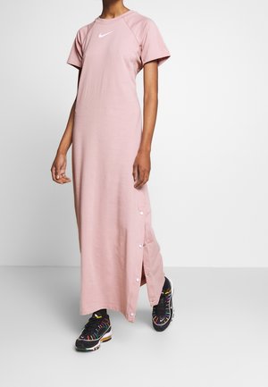 Personne portant une robe Nike rose clair, manches courtes, longueur cheville, avec des boutons-pression sur le côté, associée à des baskets noires avec des rayures colorées.