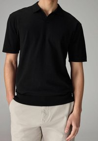 Polo noir à manches courtes avec un col en V et un col et un ourlet côtelés. Texture lisse, construction simple, associée à un pantalon beige.