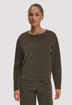 Junge Frau mit dunklem zurückgebundenem Haar, die einen passenden olivgrünen Cord-Langarmshirt und Hose trägt, steht vor einem schlichten Hintergrund.