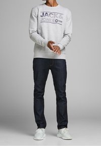 Felpa di cotone grigia chiara con testo "JACK & JONES CORE" in blu scuro, abbinata a jeans blu scuro e sneaker bianche.