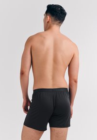 Schwarze Boxershorts mit einer weichen, glatten Textur. Der elastische Bund zeigt ein dezentes Markenlogo. Modell von hinten dargestellt.