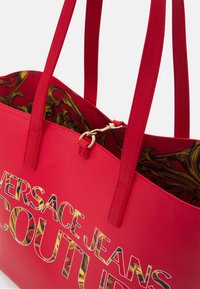 Czerwona syntetyczna torba tote z dwoma uchwytami, z złotym napisem "VERSACE JEANS COUTURE". Wnętrze podszewki wykonane z materiału w wzór.