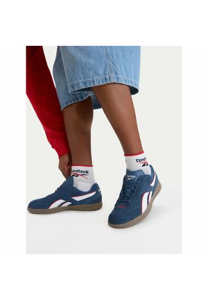 Osoba v modrých semišových teniskách s bílými a červenými detaily, s bílými ponožkami značky Reebok a džínovými šortkami, která se ohýbá, aby si upravila botu.