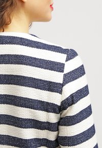 Pruhované sako s střídajícími se tmavě modrými a bílými texturovanými látkami, viditelným prošitím a na míru střiženým zadním dílem.