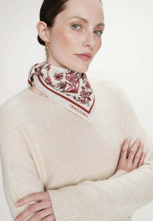 Femme à la peau claire et aux yeux bleus portant des boucles d'oreilles en or, un foulard floral autour du cou et un pull en maille couleur crème, les bras croisés.