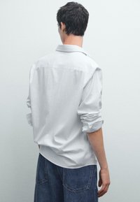 Witte, langemouwen overhemd met een button-down kraag, weergegeven vanuit de achterkant. Kenmerken zijn opgerolde mouwen en een relaxte pasvorm.