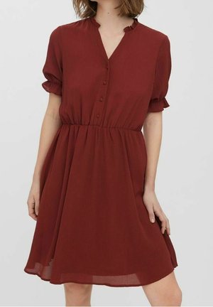 Kobieta ubrana w sukienkę do kolan w kolorze rdzawej czerwieni, z krótkim rękawem, zapinaną na guziki z przodu i elastycznym pasem, stojąca na tle jednolitego tła.