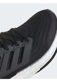 Chaussure de sport en mesh noir avec lacets noirs, support latéral en plastique et semelle en caoutchouc blanche texturée, inclinée pour montrer les détails à l'avant et sur les côtés.