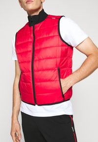 Gilet di piuma rosso con rifiniture nere, chiusura con zip frontale e tasche laterali. Presenta un colletto alto e un motivo trapuntato per un effetto di texture in più.