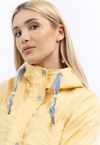 Jachetă impermeabilă galbenă cu guler înalt, dotată cu șnururi în dungi albastre și albe, accesorii din metal argintiu și o textură netedă.