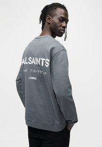 Sweat-shirt gris avec "ALLSAINTS" en grandes lettres blanches dans le dos, accompagné de textes supplémentaires en plusieurs langues. Matière en coton doux.