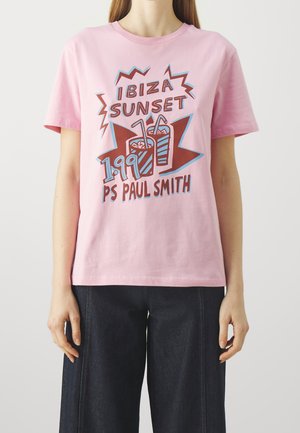 Frau trägt ein pinkes T-Shirt mit grafischem Text "Ibiza Sunset 1991 PS Paul Smith" und zwei illustrierten Getränken auf der Vorderseite.