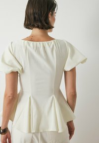 Top peplo bianco con maniche corte a sbuffo, tessuto testurizzato e scollatura leggermente arricciata. Il retro presenta cuciture verticali e un orlo svasato.