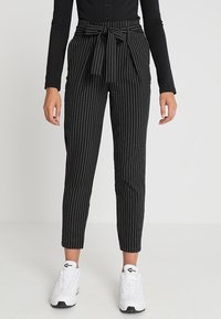 Black high-waist pinstripe pants with front tie bow worn with black long-sleeve top and white sneakers on plain background.