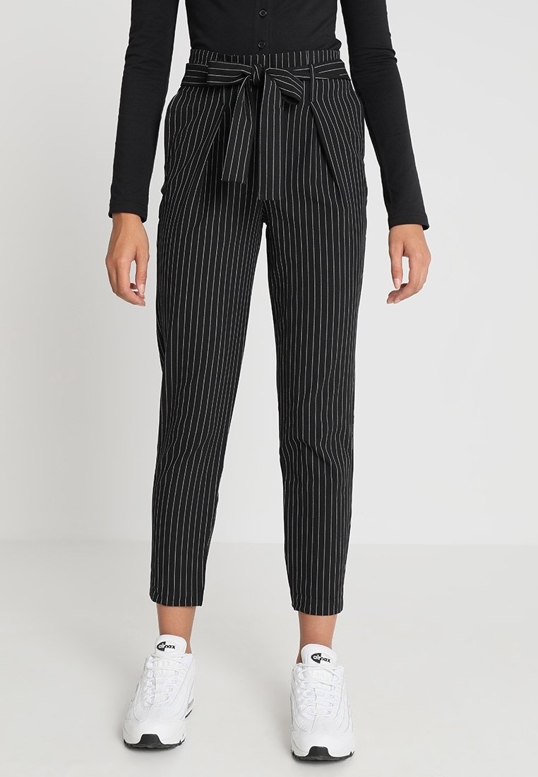 Black high-waist pinstripe pants with front tie bow worn with black long-sleeve top and white sneakers on plain background.