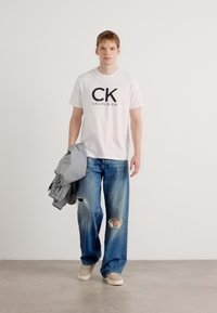 Witte katoenen t-shirt met zwarte "CK CALVIN KLEIN" logo, gecombineerd met blauwe versleten jeans met grote scheuren en beige sneakers. Grijze jas in de hand.