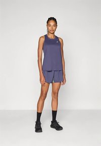 Mlada žena stoji u ljubičastom sportskom topu i šorcu, s crnim čarapama i tenisicama, nasuprot jednostavne pozadine.