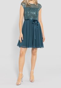 Robe teal avec un corsage en sequins, jupe lisse avec plis et ceinture en satin. Portée avec des sandales à talons transparents.