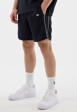 Uomo che indossa pantaloncini blu navy con bordi bianchi, calzini bianchi alla caviglia e scarpe da ginnastica bianche, in piedi contro uno sfondo chiaro e semplice.