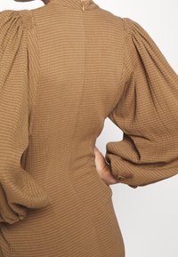 Mujer vistiendo un vestido marrón texturizado con mangas largas abullonadas y un cierre en la parte posterior, con la mano colocada en la cintura.