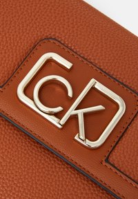 Hnědá texturovaná kožená kabelka s výrazným zlatým logem "CK" na obdélníkové nášivce. Hladké okraje s akcenty švu.