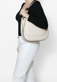 Borsa a tracolla in pelle beige con forma curva, bordature nere e un accento dorato. Indossata sulla spalla, abbinata a pantaloni bianchi.