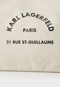 Beiger Canvas-Shopper mit schwarzem Drucktext: "KARL LAGERFELD," "PARIS," und "21 RUE ST-GUILLAUME." Schlichte, rechteckige Form.