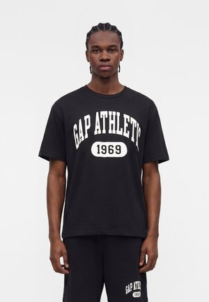 Νεαρός με πλεξούδες, ντυμένος με μαύρο T-shirt GAP ATHLETICS 1969 και αντίστοιχα σορτς, στέκεται μπροστά σε απλό φωτεινό φόντο.