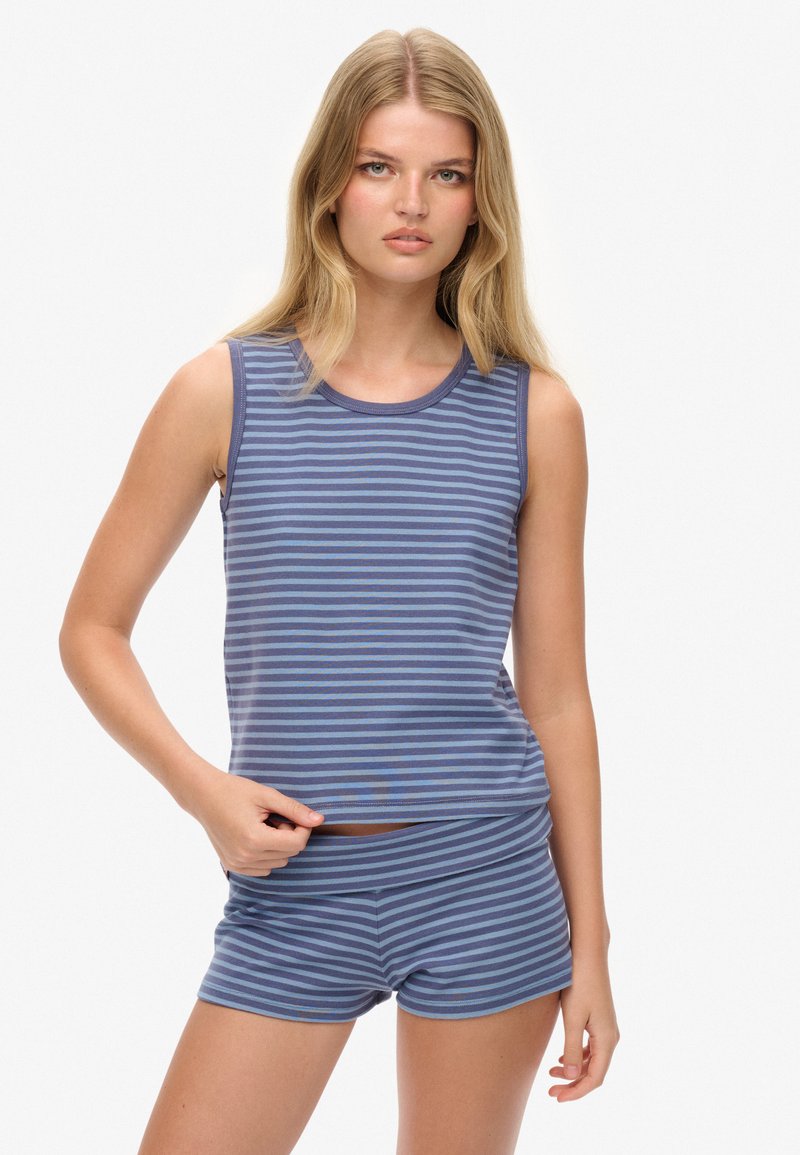 Jonge vrouw met lang blond haar, die een blauw en grijs horizontaal gestreept mouwloos topje en bijpassende shorts draagt, staand tegen een witte achtergrond.