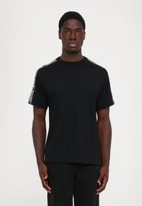 Άνδρας φορά μαύρη μπλούζα με λευκές διακριτικές ρίγες στους ώμους και μαύρο παντελόνι, στέκεται μπροστά σε ένα λευκό φόντο.