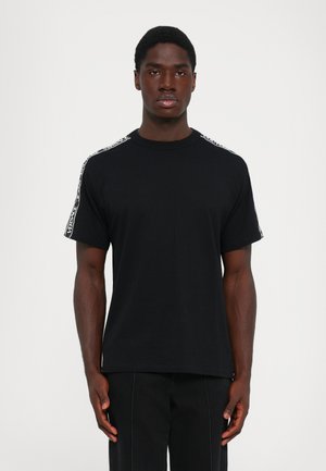 Man wearing a black t-shirt with white branded stripes on the shoulders and black pants, standing against a plain white background.