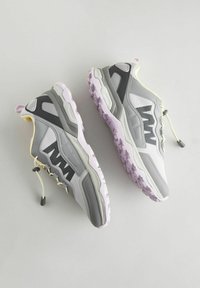 Baskets grises et blanches avec un extérieur texturé, dotées d'une semelle violette et d'un système de lacets élastiques. Des panneaux latéraux renforcés offrent un soutien.