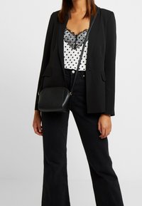 Svart kavaj, vit blus med svarta prickar och spetsdetaljer, utställda svarta jeans och en liten svart axelväska med dragkedja.