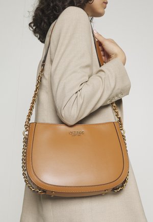 Vrouw met beige blazer houdt een tan leren Guess schoudertas met goudkleurige kettingriem vast tegen een lichte achtergrond.