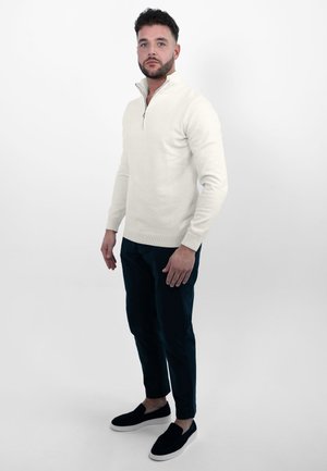 Lichtcrème zip-kraag trui, slim-fit donkere broek en zwarte instapschoenen. Gladde textuur, minimale accenten en een ontspannen ontwerp.