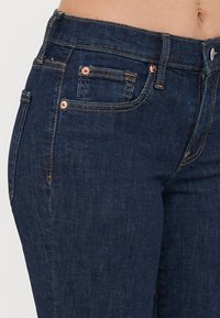 Jean en denim bleu foncé avec une coupe slim, des rivets en cuivre et des coutures contrastées. Le tissu a une texture lisse et peu d'ornements.