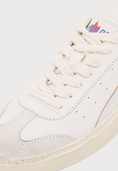 Gros plan d’une basket blanche avec des lacets texturés, un bout en suède et un logo partiel sur la languette sur un fond clair.