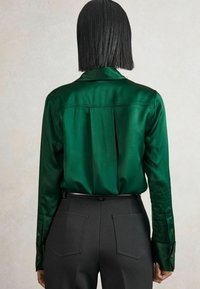 Person mit glattem schwarzen Haar, der ein glänzendes grünes Langarmshirt trägt, das in hochgeschnittene schwarze Hosen mit Gesäßtaschen gesteckt ist.