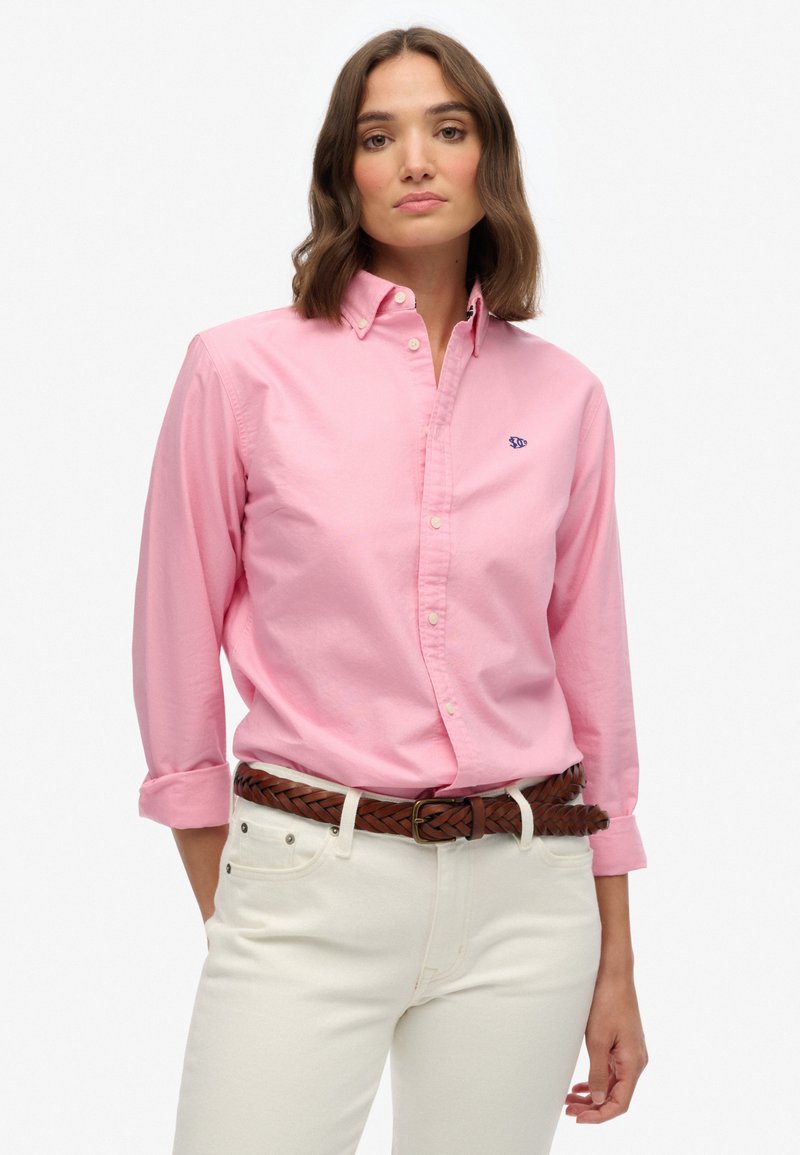 Lichtroze shirt met knopen en lange mouwen, met een kraag en geborduurd logo. Gedragen met een gevlochten bruine riem en een witte broek.