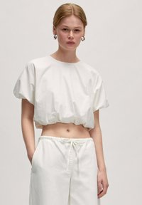 Witte cropped top met brede mouwen en een gerimpelde onderrand, gemaakt van een lichte, gladde stof. Gecombineerd met witte joggingbroek met trekkoord.