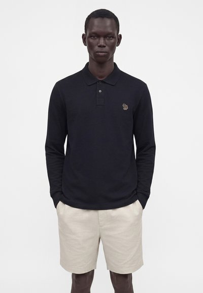 Homme portant un polo noir à manches longues avec un petit logo zèbre et un short beige, debout, les mains dans les poches, sur fond blanc.