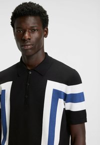 Ung mand med kort krøllet hår iført en sort polo skjorte med hvide og blå geometriske striber på en ensfarvet baggrund.