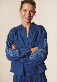 Denim-paita pitkillä hihoilla, käännettävällä kauluksella ja kukkakirjontaa hihoissa. Sininen väri, rento malli ja kerättyjä yksityiskohtia.