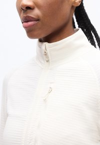 Κοντινό πλάνο ενός ατόμου που φοράει άσπρο πλεκτό πουλόβερ με φερμουάρ, όρθιο γιακά και τα μαλλιά του είναι πλεγμένα και δεμένα πίσω.