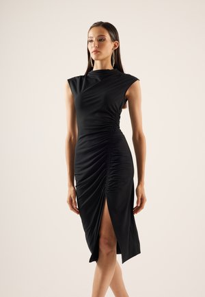 Vrouw draagt een mouwloze zwarte geplooide jurk met een zijsplit, staat met ontspannen armen tegen een effen beige achtergrond.