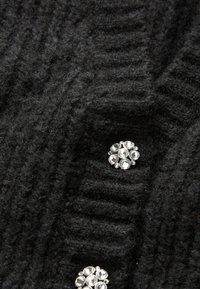 Μαύρος πλεκτός ύφασμα με ριχτές υφές, που διαθέτει μια διακοσμητική ασημένια καρφίτσα με πολλές διάφανες πέτρες τοποθετημένες σε κυκλικό σχέδιο.