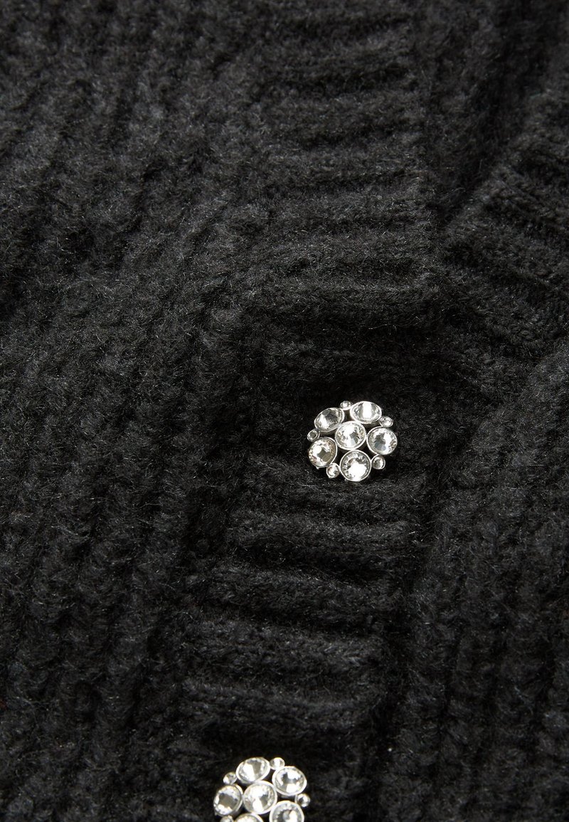 Czarna dzianina o prążkowanej fakturze, zdobiona dekoracyjną srebrną broszką z wieloma przezroczystymi kamieniami ułożonymi w okrągły wzór.